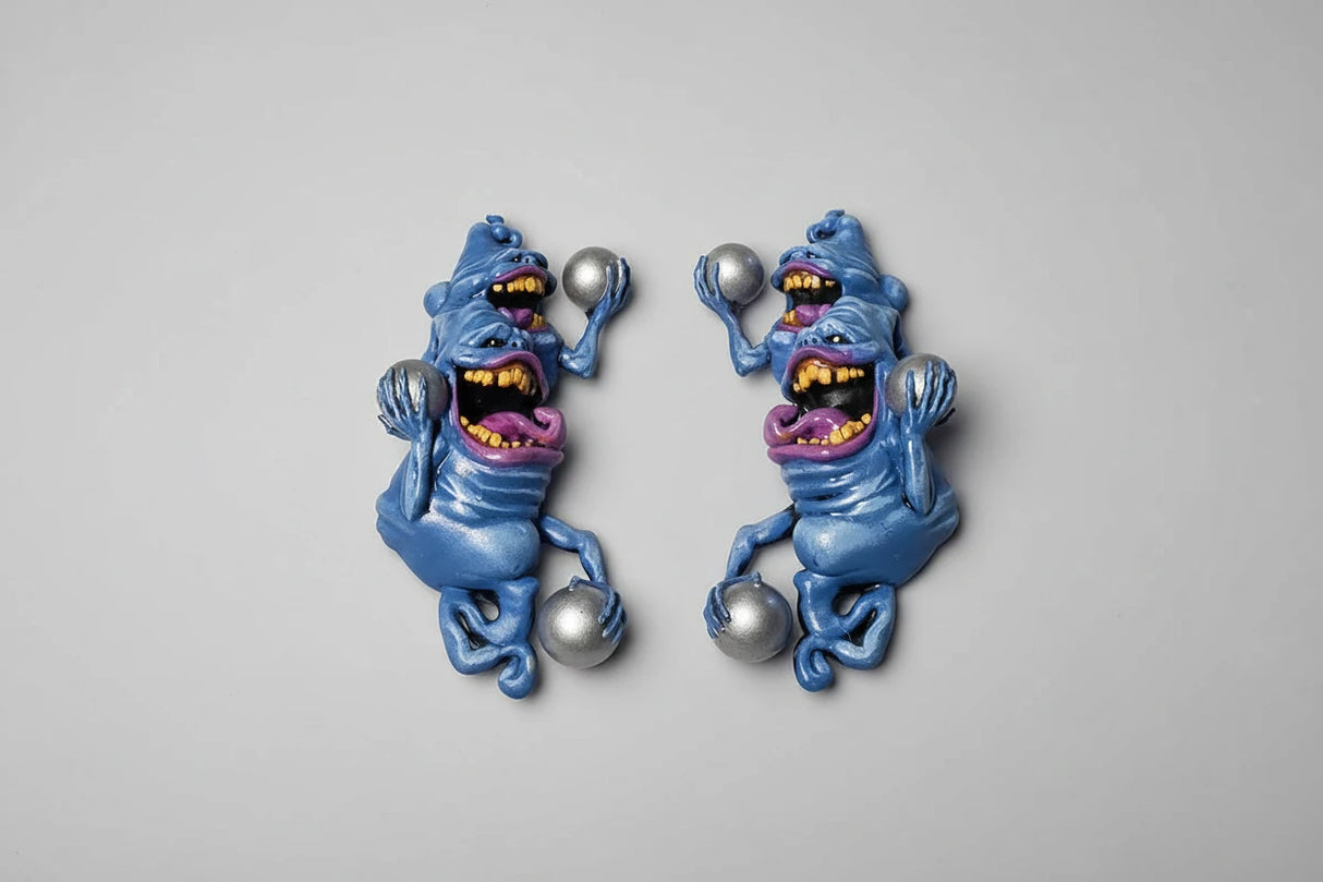 Pair of blue creature sculpts with yellow mouths and purple tongues holding pinballs on a gray background
