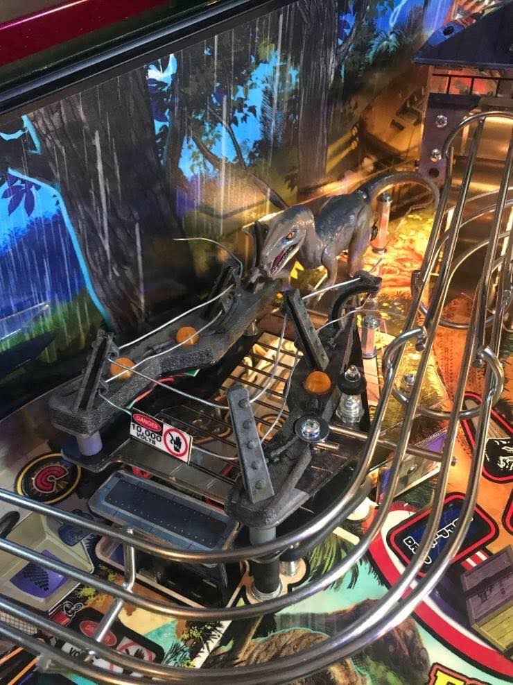 Realistic-looking raptor pen installed on a pinball machine playfield, with orange domes and a '10,000 volts' sign.