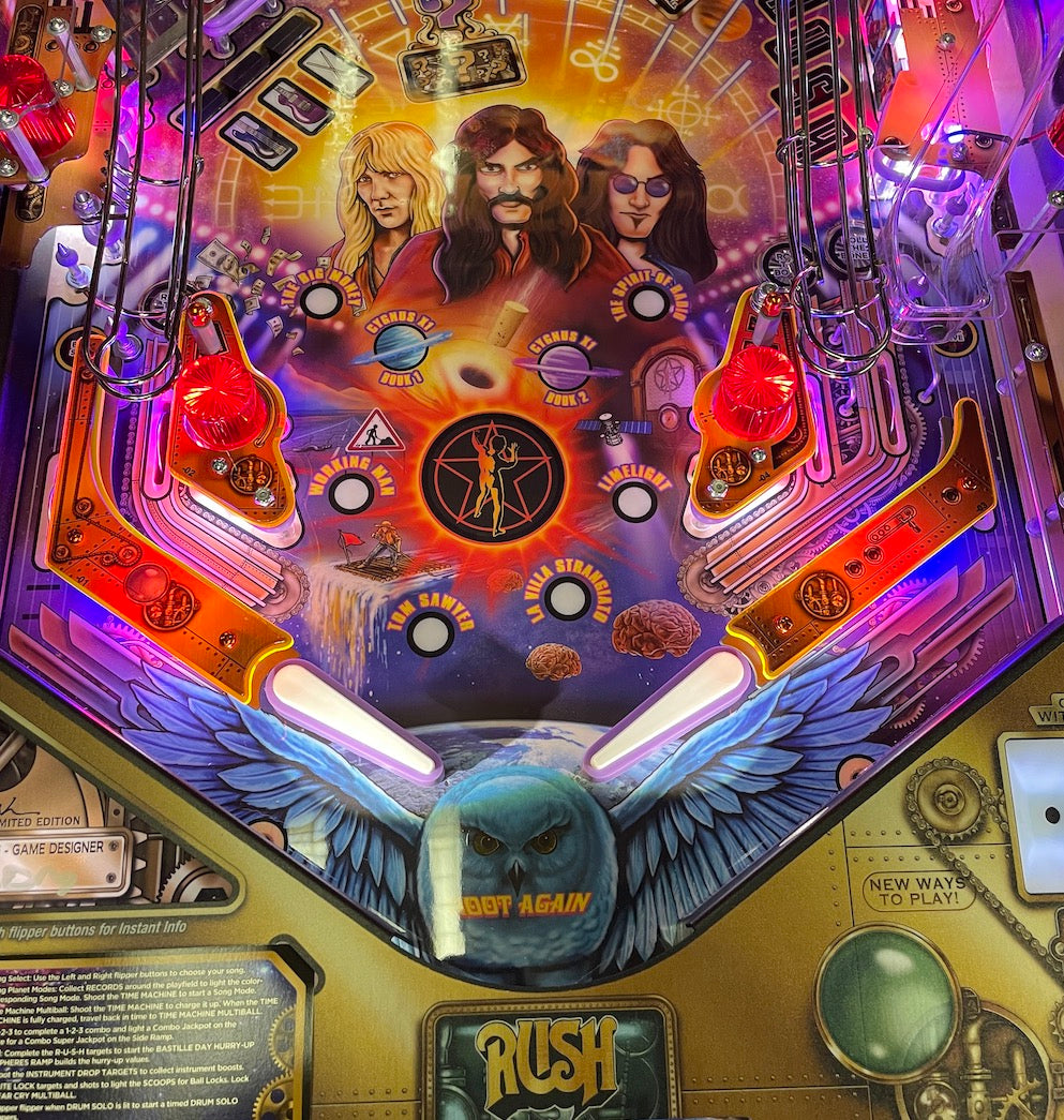 Close-up view of a pinball machine's playfield featuring colorful artwork with Rush band members and celestial motifs, highlighting the plastic protector set installed around the game's hardware.