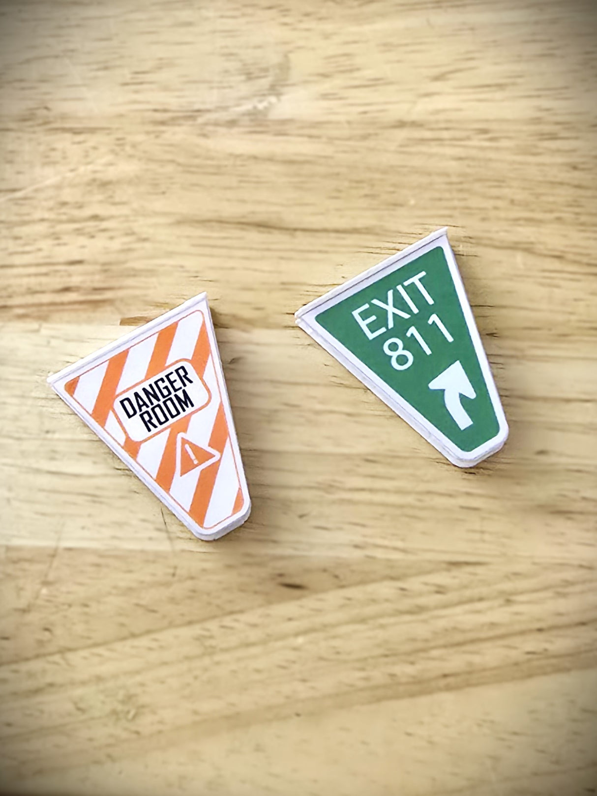 Two pinball machine gate covers with 'Uncanny X-Men' themed designs, one orange with 'DANGER ROOM' text and one green with 'EXIT 817' text on a wooden surface.