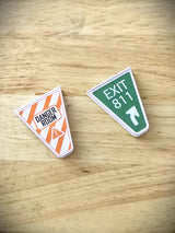 Two pinball machine gate covers with 'Uncanny X-Men' themed designs, one orange with 'DANGER ROOM' text and one green with 'EXIT 817' text on a wooden surface.