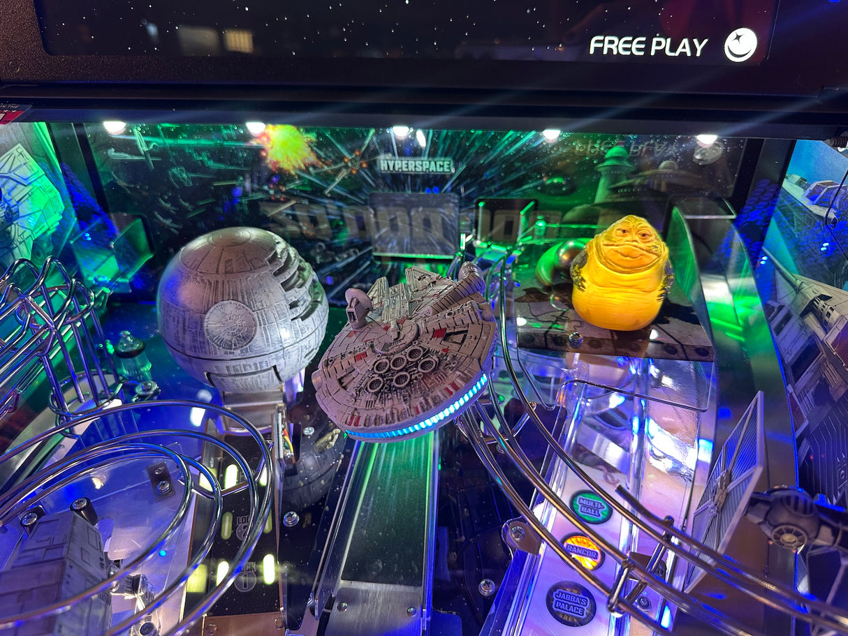 Pinball machine with Star Wars-themed decorations, including a Death Star and Millennium Falcon, in a dark room.