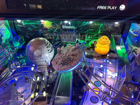 Pinball machine with Star Wars-themed decorations, including a Death Star and Millennium Falcon, in a dark room.